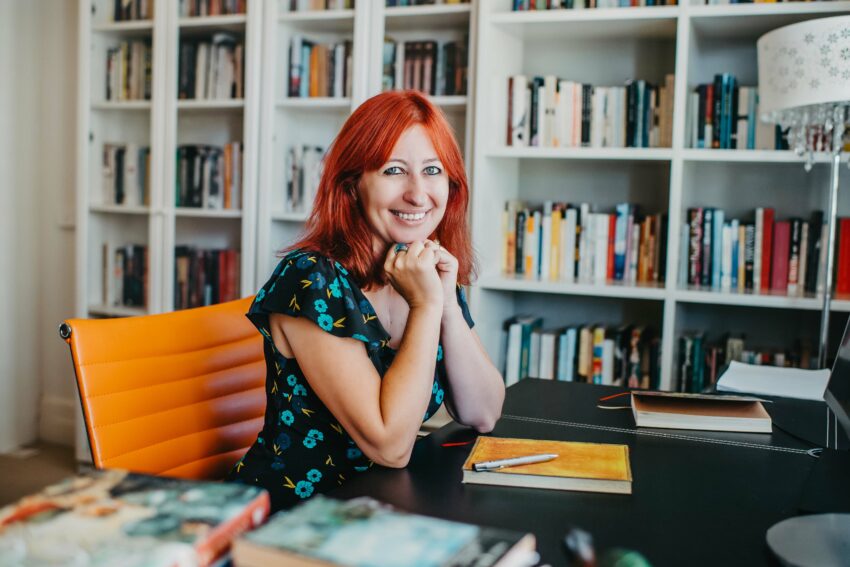Image of writer Lee Kofman at her desk in front of book shelves.
